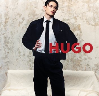 Joven con chaqueta negra, camisa blanca con rayas y corbata negra, de pie con las manos en las caderas frente a una pared texturizada y un sofá cubierto, texto "HUGO" en rojo.