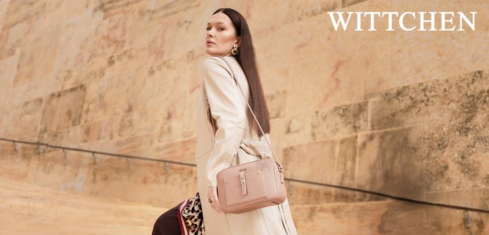 Frau im beigefarbenen Mantel posiert mit rosa Handtasche vor einer strukturierten Steinmauer, elegant und stilvoll mit langen dunklen Haaren.