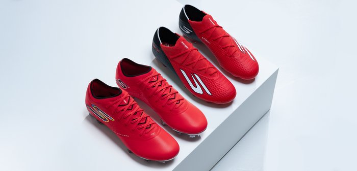 Τέσσερα κόκκινα ποδοσφαιρικά παπούτσια με μαύρες και λευκές λεπτομέρειες, τακτοποιημένα σε δύο ζευγάρια πάνω σε μια ανυψωμένη λευκή πλατφόρμα με φόντο ανοιχτόχρωμο.