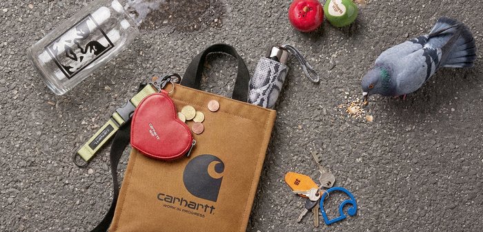 Sac Carhartt marron sur le trottoir avec des pièces de monnaie, une pochette rouge en forme de cœur, des clés, une bouteille d'eau, un parapluie, deux pommes et un pigeon mangeant des miettes.