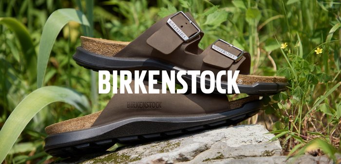 Zwei braune Birkenstock-Sandalen mit Schnallenriemen im Freien auf Felsen gestapelt, umgeben von grünen Pflanzen.