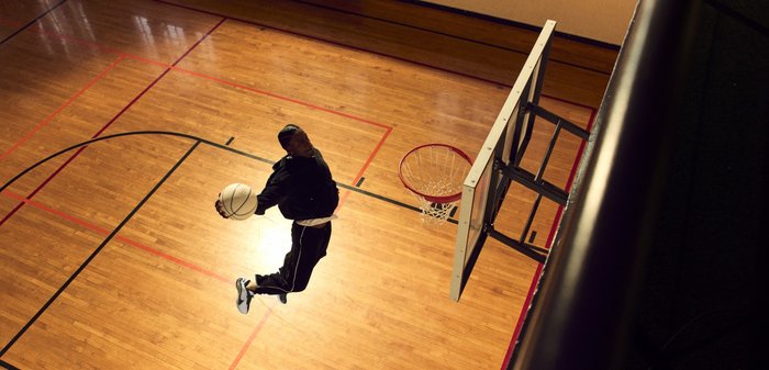 Joueur de basketball en plein saut effectuant un dunk avec un ballon de basket blanc. Il porte un survêtement noir avec des accents blancs, sur un parquet en bois aux lignes rouges.