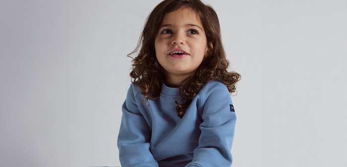 Jong meisje met krullend bruin haar, gekleed in een blauwe trui, kijkt omhoog en lacht tegen een effen grijze achtergrond.