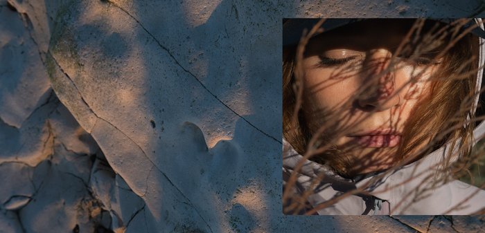 Woman with closed eyes wearing a light jacket, shadows of bare branches cast on her face, against a textured rock background.