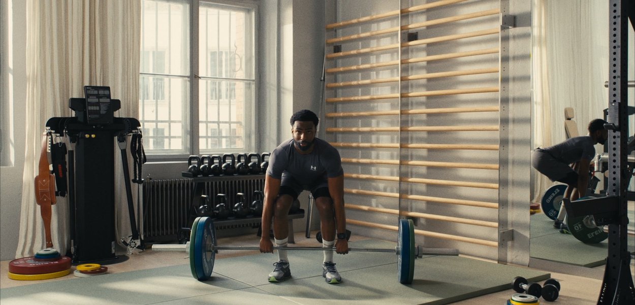 Hombre con ropa deportiva levantando una barra en un gimnasio con pesas, kettlebells y equipo de ejercicio cerca de grandes ventanas.