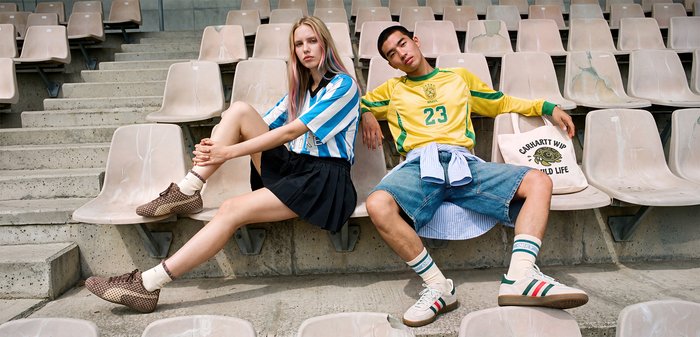 Deux jeunes adultes en maillots de sport assis décontractés dans des sièges vides du stade, une femme blonde avec une jupe noire, un homme en bermuda en jean.