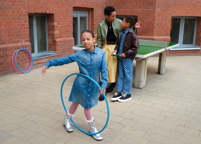 Une fille en robe bleue joue avec un cerceau bleu pendant qu'une femme et un garçon se tiennent à proximité près d'une table de ping-pong en extérieur.