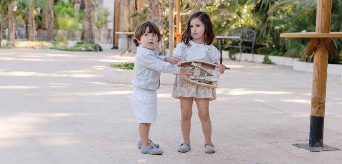Two young children stand outdoors, one handing a small wooden boat with a net to the other, both wearing light-colored clothes and shoes.
