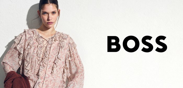 Young woman wearing a patterned blouse and jeans, holding a knitted sweater, standing against a white wall with the word "BOSS" overlayed.