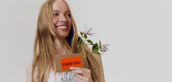 Femme souriante aux longs cheveux tenant des fleurs violettes et une carte cadeau Zalando orange sur un fond clair uni.