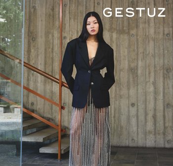 Crni veliki sako s jednim gumbom, upareni s prozirnom haljinom s vertikalnim uzorcima od perlica, postavljen pred teksturiranim zidom.