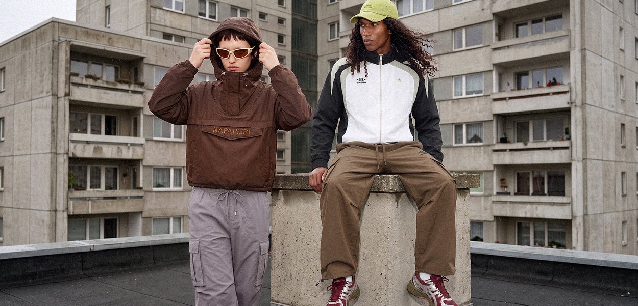 Dos adultos jóvenes vestidos con ropa informal de calle, uno ajustándose las gafas de sol, el otro sentado en el borde de hormigón con edificios de apartamentos al fondo.