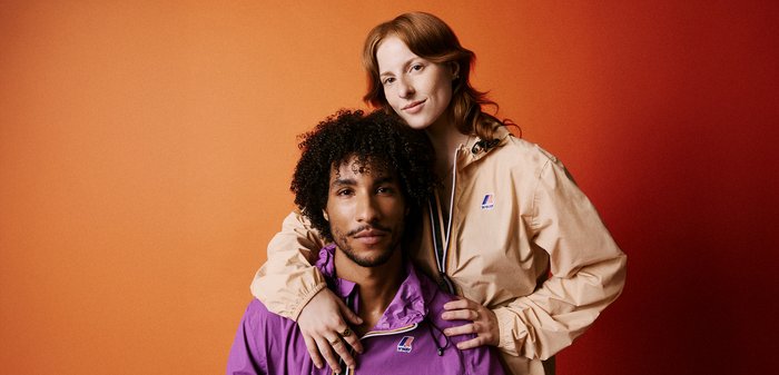 Leggeri giacconi viola e beige, con dettagli a zip e cordini, indossati da due persone su uno sfondo arancione sfumato.