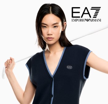 Młoda kobieta w granatowej bezrękawnikowej dzianinowej kamizelce trzyma białą kij golfowy za ramionami, z logo EA7 Emporio Armani w prawym górnym rogu.