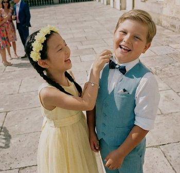 Ragazza in vestito giallo e fascia floreale sistema la papillon di un ragazzo; entrambi sorridono all'aperto su un pavimento in pietra durante un evento formale.