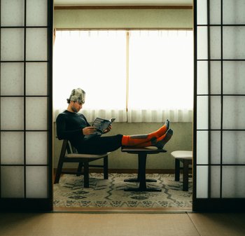 Schwarzes Langarm-Oberteil und orangefarbene Socken. Person sitzt auf einem Holzstuhl an einem niedrigen Tisch und liest ein Buch, wobei Licht durch transparente Vorhänge fällt.