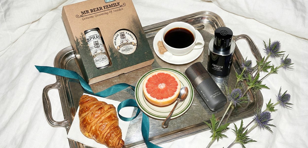 Bandeja de plata que sostiene una taza de café negro, medio pomelo en un plato, un cruasán, productos de cuidado personal y flores de cardo púrpura sobre tela blanca.