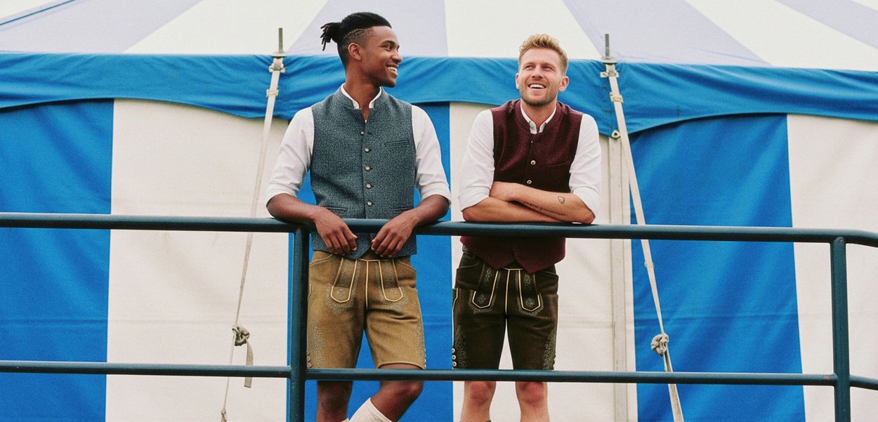 Two people wear traditional attire: one in a blue-gray vest and shorts, the other in a burgundy vest and dark shorts. Both have white shirts.