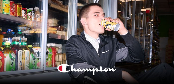 Young man in Champion tracksuit drinking from a can while sitting by a refrigerated beverage display in a store aisle.