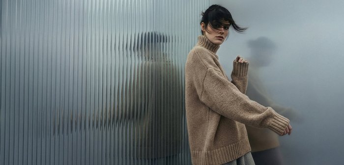 Γυναίκα με μπεζ πουλόβερ με ζιβάγκο, τα μαλλιά της ανεμίζουν, στέκεται δίπλα σε γυάλινο τοίχο με υφή που αντανακλά το σιλουέτα της.