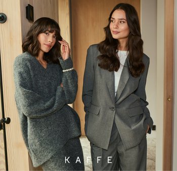 Twee vrouwen staan in een moderne interieur. De ene draagt een grijze, oversized, gestructureerde trui; de andere een getailleerd grijs streepjespak met een witte top.