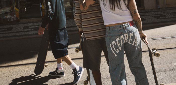 Three tattooed young people holding skateboards, standing on a sunlit street with tram tracks visible.