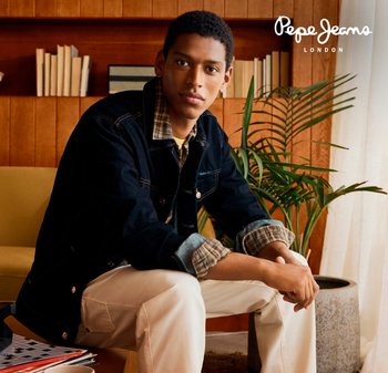 Joven en una chaqueta de mezclilla oscura y pantalones beige, sentado en un sofá amarillo en una habitación de madera con libros y una gran planta enmacetada.