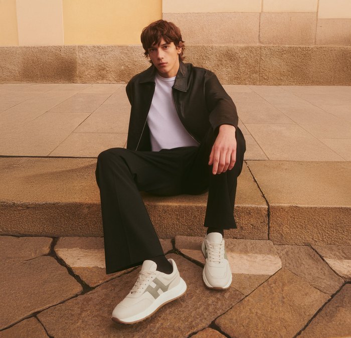 Giovane uomo seduto su gradini di pietra, indossa giacca di pelle nera, camicia bianca, pantaloni neri e sneakers bianche con logo "H".