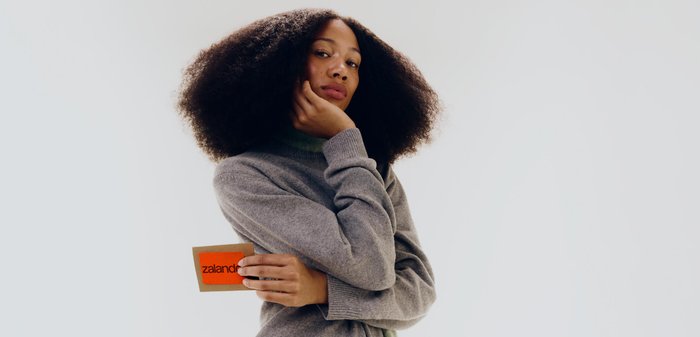 Frau mit natürlich lockigem Haar, die einen grauen Pullover trägt, hält eine orangefarbene Karte mit dem Text "zalando" vor einem einfarbigen weißen Hintergrund.