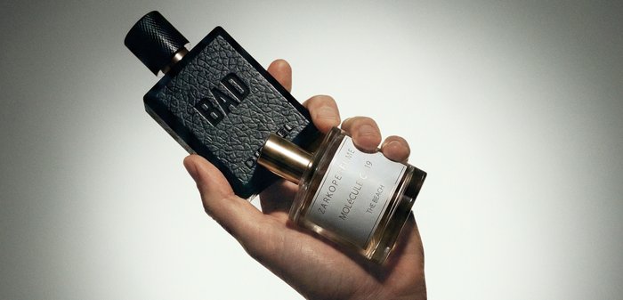 Main tenant deux flacons de parfum, l'un noir texturé avec l'étiquette "BAD", l'autre transparent avec une étiquette blanche portant l'inscription "Zarkoperfume Molecule 19".