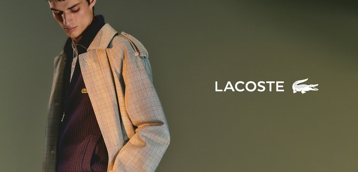 Manteau à carreaux en tons beige et bleu, avec une fermeture à boutons sur le devant, et un look décontracté superposé avec un pull bleu marine en dessous. Logo LACOSTE visible.