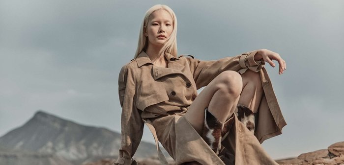 Femme aux longs cheveux blonds portant un trench-coat beige et des bottes en peau de vache, assise sur un terrain rocheux sous un ciel nuageux.
