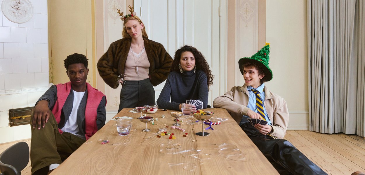 Keturi jauni suaugusieji sėdi ir stovi aplink medinį stalą su kortomis, kauliukais, saldainiais ir šventinėmis dekoracijomis jaukiame kambaryje.