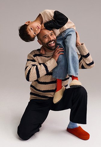 Homme à genoux, souriant, tenant de manière ludique un enfant la tête en bas sur ses épaules, tous deux portant des vêtements décontractés et des chaussettes colorées.