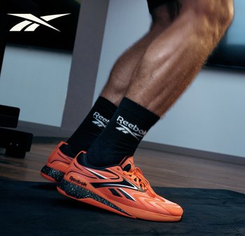 Person mit leuchtend orangefarbenen Reebok-Schuhen und schwarzen Reebok-Socken, stehend auf einer dunklen Bodenmatte in einem Fitnessstudio.