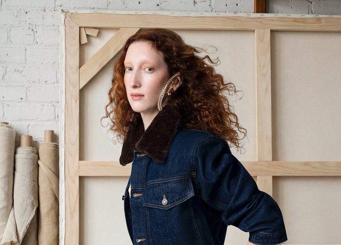 Γυναίκα με κόκκινα μαλλιά και μεγάλο χρυσό κρίκο στο αυτί φοράει σκούρο τζιν μπουφάν με γούνινο γιακά, στέκεται μπροστά σε ξύλινα πλαίσια καμβά και τυλιγμένο ύφασμα.
