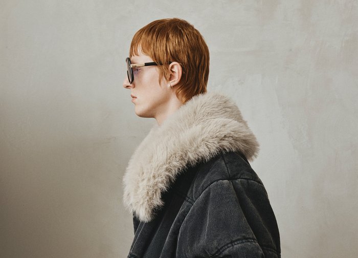 Veste en velours côtelé noire avec un large col en fausse fourrure gris clair. Modèle portant de grandes lunettes de soleil, de profil, sur un fond uni.