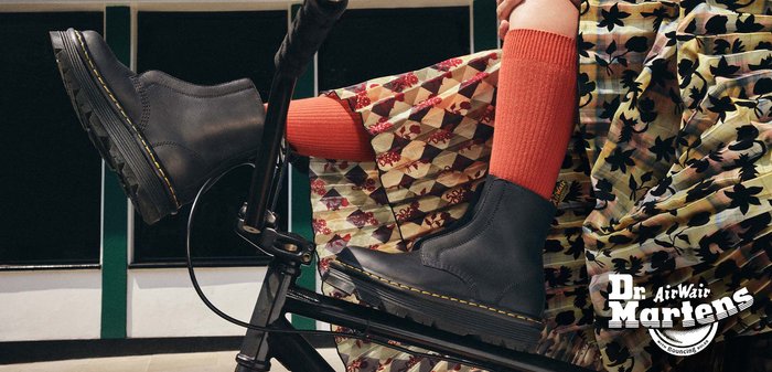 Botines de cuero negro hasta el tobillo con un acabado suave, costuras amarillas y una suela gruesa y texturizada, combinados con calcetines naranjas de canalé y una prenda con estampado.