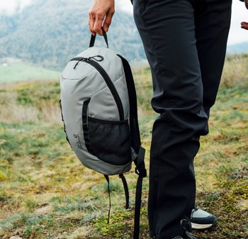 Grauem Rucksack mit runder Form, der eine seitliche Netztasche und einen oberen Griff hat, wird an der Seite einer Person in schwarzer Hose gehalten.