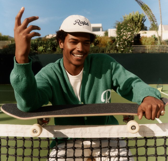 Ung mand smilende, iført grøn cardigan og hvid kasket, holder skateboard over et tennisnet på en udendørs bane.