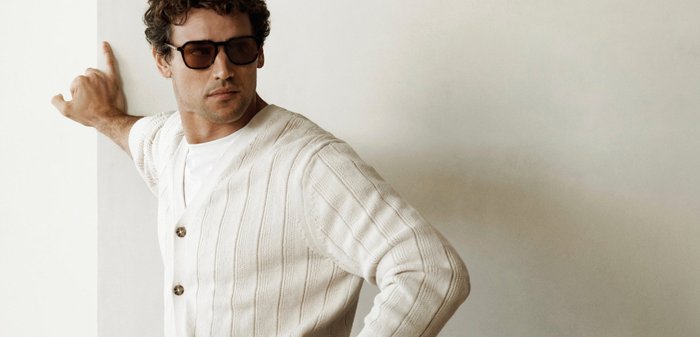 Vyras dėvi rudus saulės akinius, baltą kardiganą ir laikrodį, pozuoja viena ranka ant klubų, kita ant sienos, paprastas fonas.