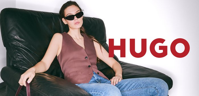Brauner ärmelloser Weste mit Knöpfen, blaue Jeans und schwarze Sonnenbrille, während sie auf einem schwarzen Lederstuhl sitzt; roter Text "HUGO" daneben.
