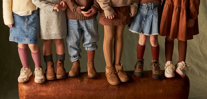 Hohe Sneaker aus elfenbeinfarbenem Leder mit braunen Gummisohlen, ausgestattet mit einem Klettverschluss. Dunkel gerippte Strumpfhosen und ein lila Strickoberteil sind sichtbar.