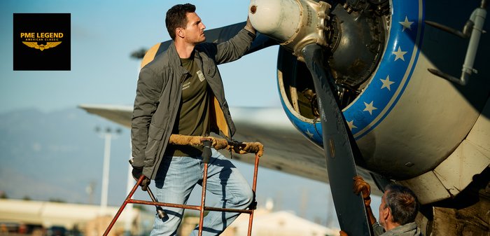 Man in jas en spijkerbroek inspecteert buiten een vintage vliegtuigpropeller met een andere persoon, bij het PME Legend-logo.