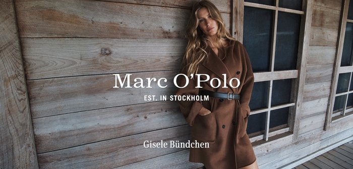 Brązowy wełniany płaszcz typu trench z podwójnym zapięciem, dużymi kieszeniami i czarnym paskiem, umieszczony na tle drewnianej ściany z oknami.