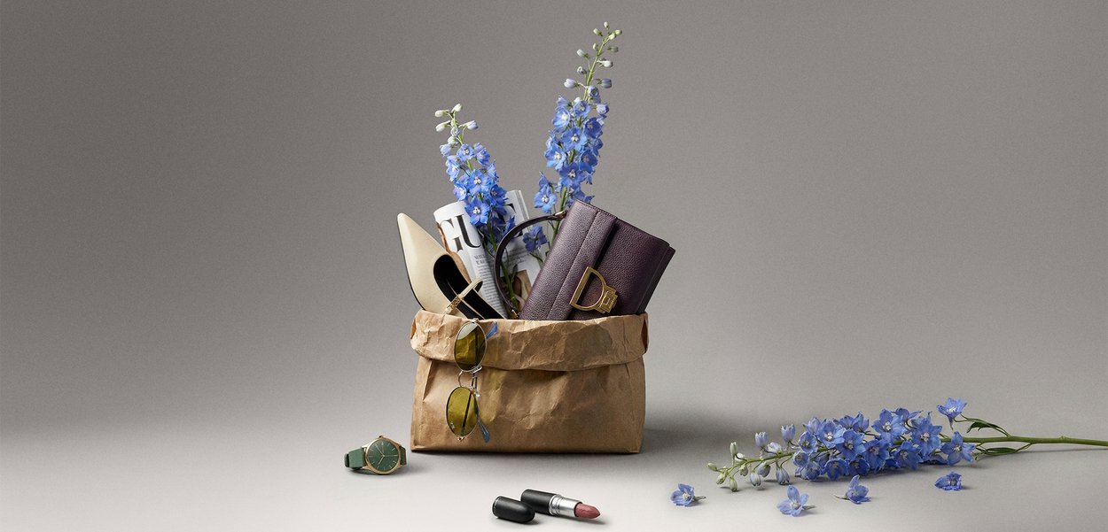 Ruskea paperipussi, jossa on kerma valkoinen korkokenkä, violetti kirjekuorilaukku, rullattu aikakauslehti, aurinkolasit ja sinisiä kukkia, lähellä punainen huulipuna ja kello.