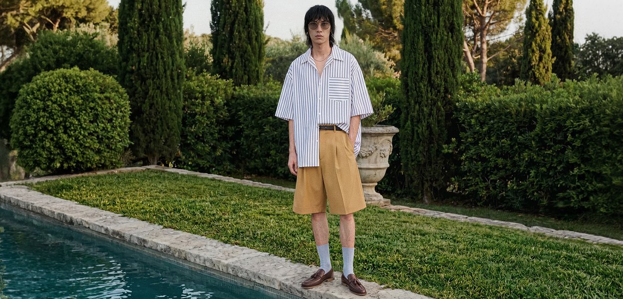 Persona con camisa de manga corta a rayas oversized, pantalones cortos mostaza, calcetines azul claro y zapatos marrones, de pie junto a una piscina con vegetación de fondo.