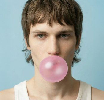 Ung person med kort brunt hår som bär en vit linne och blåser en stor rosa tuggummibubbla mot en ljusblå bakgrund.