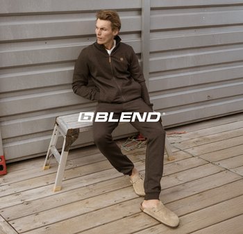Felpa in pile marrone scuro con zip e un piccolo logo, abbinata a pantaloni jogger coordinati e scarpe slip-on beige, su una superficie di legno.