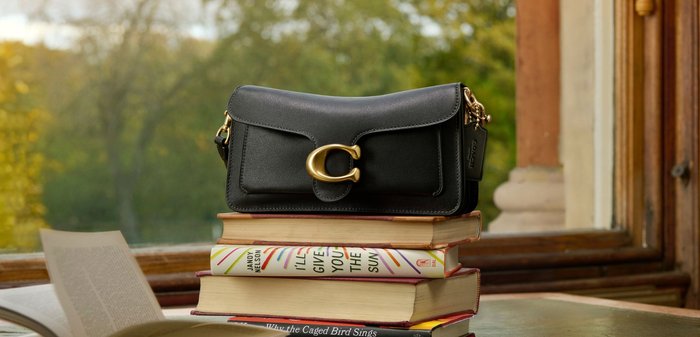 Schwarze Lederhandtasche mit goldener Schnalle liegt auf einem Stapel von vier Büchern neben einem aufgeschlagenen Buch auf einem Holztisch am Fenster.